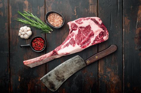 Raw uncooked black angus beef tomahawk steak bone set old butcher cleaver knife with seasoning herbs old dark wooden table